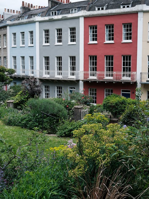 A small urban garden situated in front of a multi-storey building painted in pastel blue and pink with white window frames and a narrow white balcony running across the second floor. The garden featur