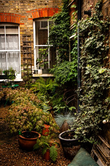 A small urban garden situated in front of a multi-storey building painted in pastel blue and pink with white window frames and a narrow white balcony running across the second floor. The garden featur