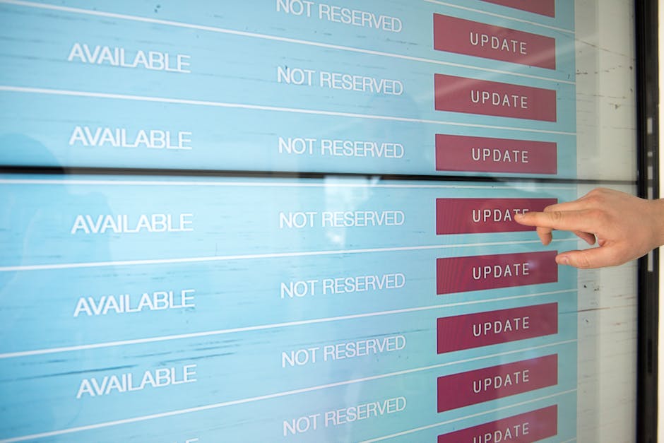 A person’s hand with neatly trimmed nails and light skin tone is pointing to an available slot on a digital booking screen that displays a list of time slots. The screen has entries labeled 'AVAILABLE
